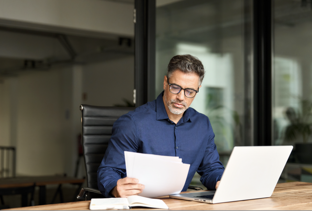 Senior mature male account bank manager executive working on laptop checking paper financial report in office. Busy middle aged professional business man using laptop computer reading document at work See Less By insta_photos